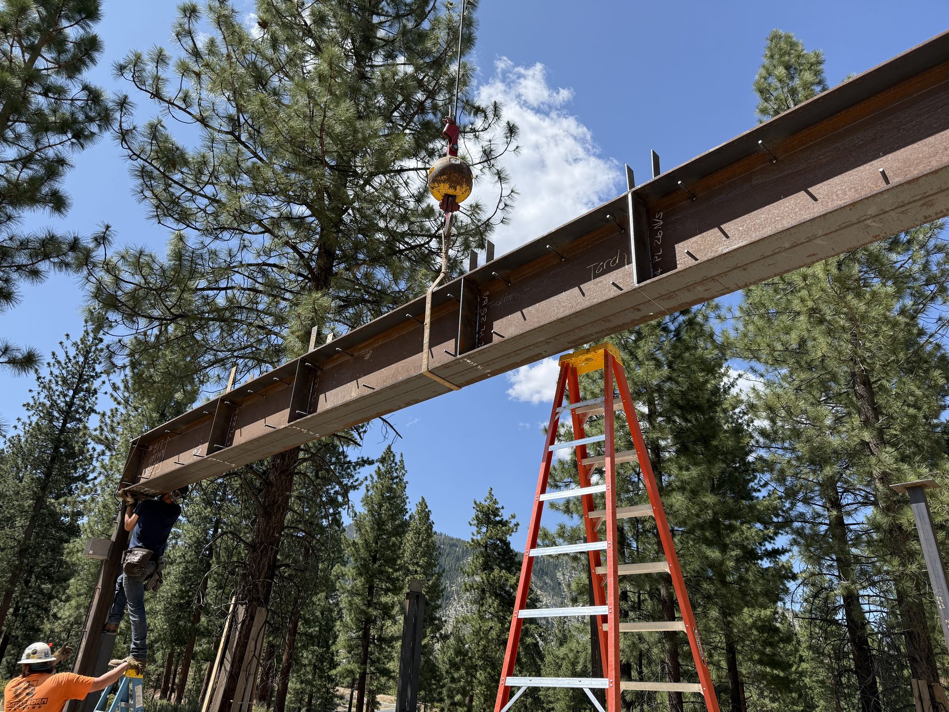 Steel beam being positioned by crane with worker on ladder below
